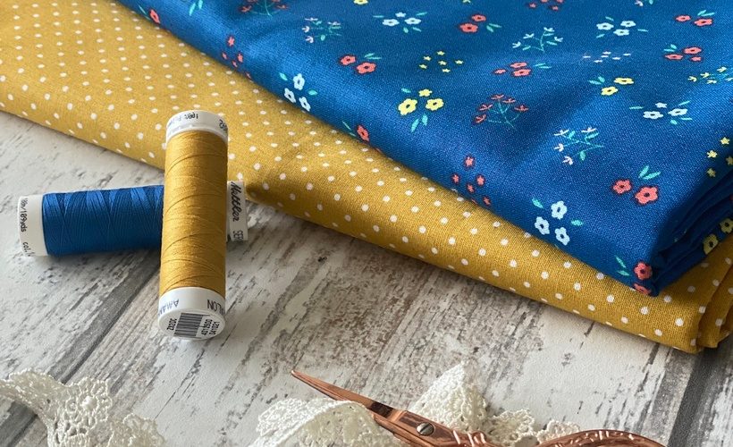 blue floral fabric sitting on top of yellow polka dot fabric with blue and yellow cotton reels lace and rose gold scissors